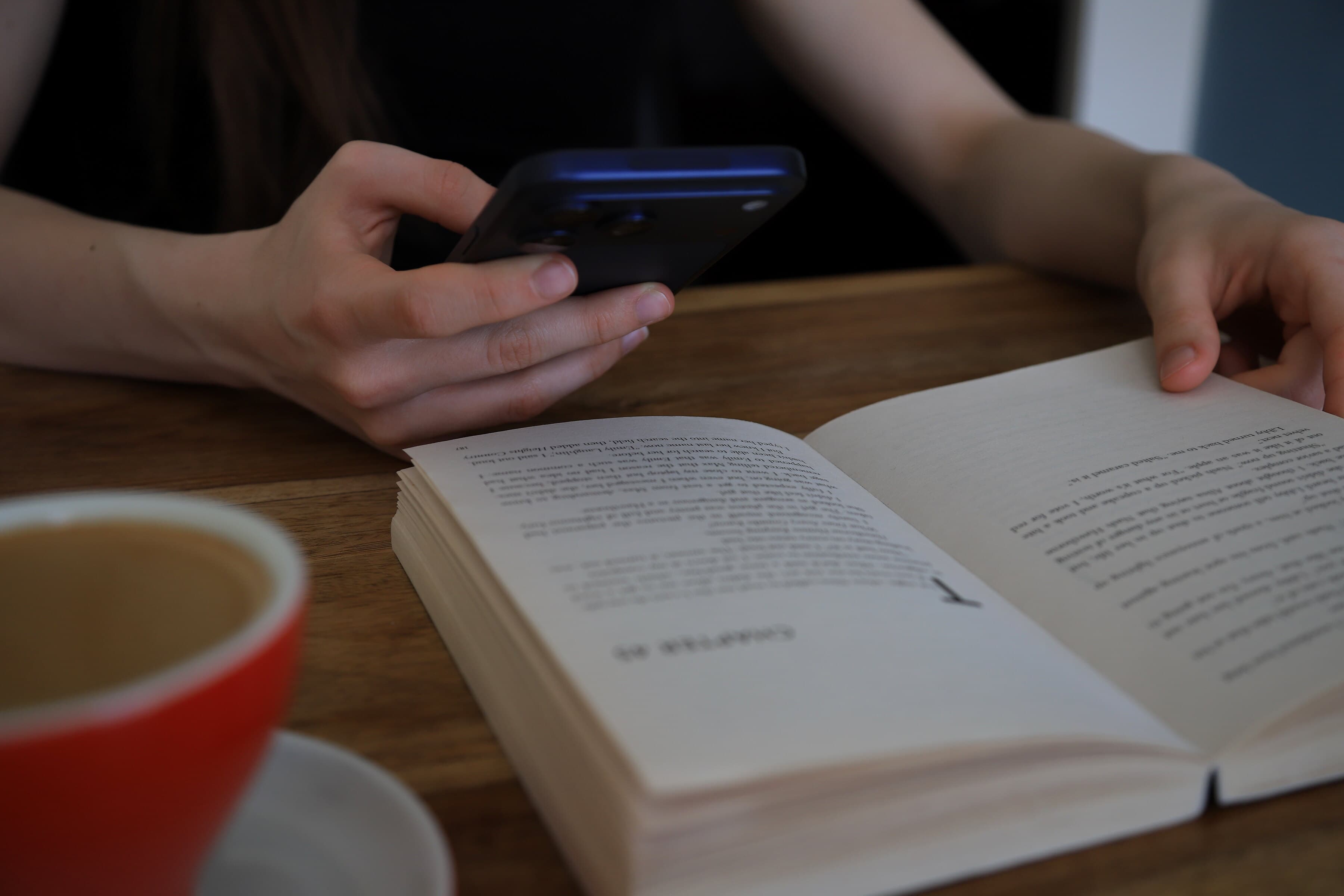 A reader in a dark top holds an iPhone aimed at an open novel on a wooden table, a red cup in the foreground